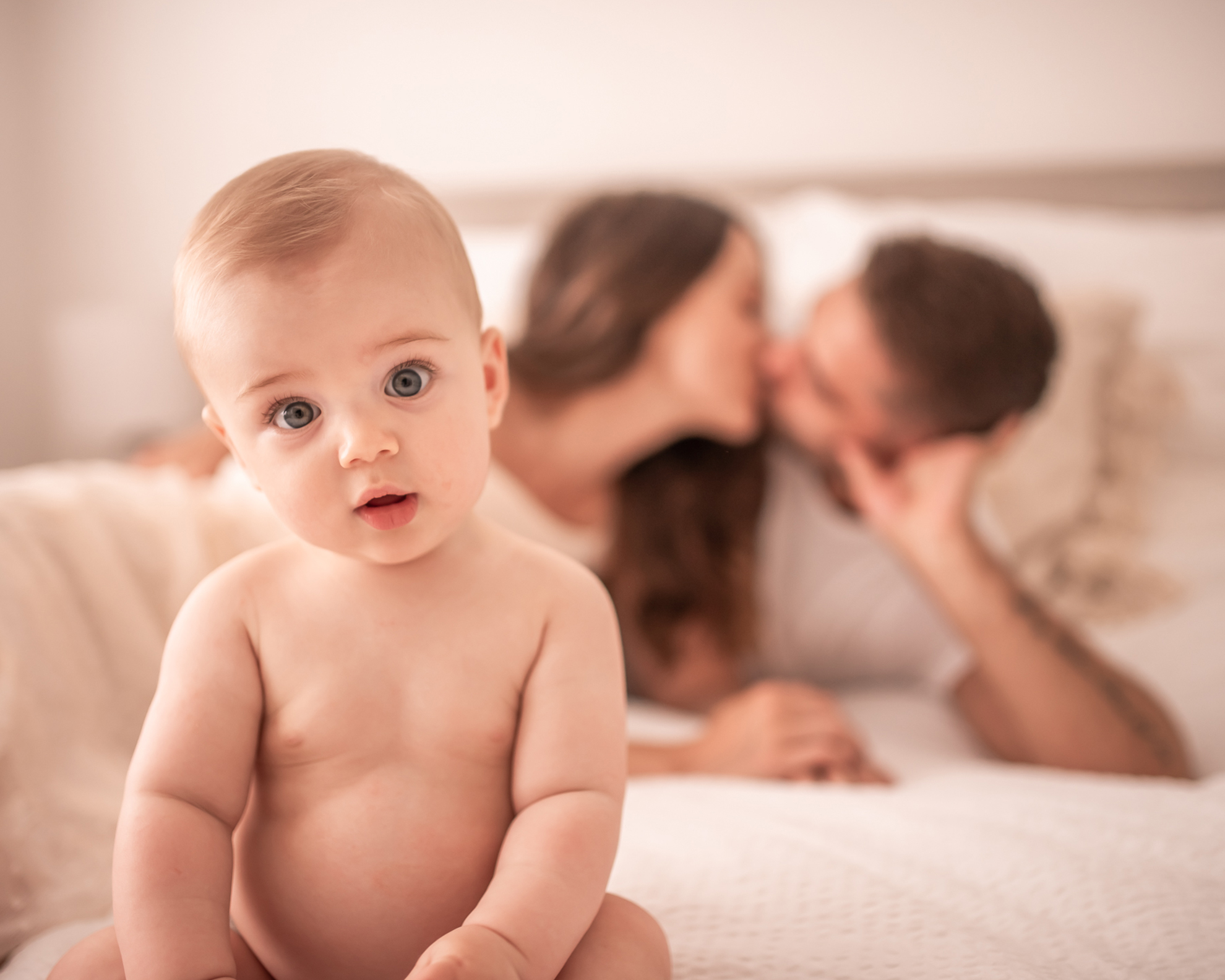 portrait of a baby in the foreground and his parents kissing in the background during at home family photoshoot